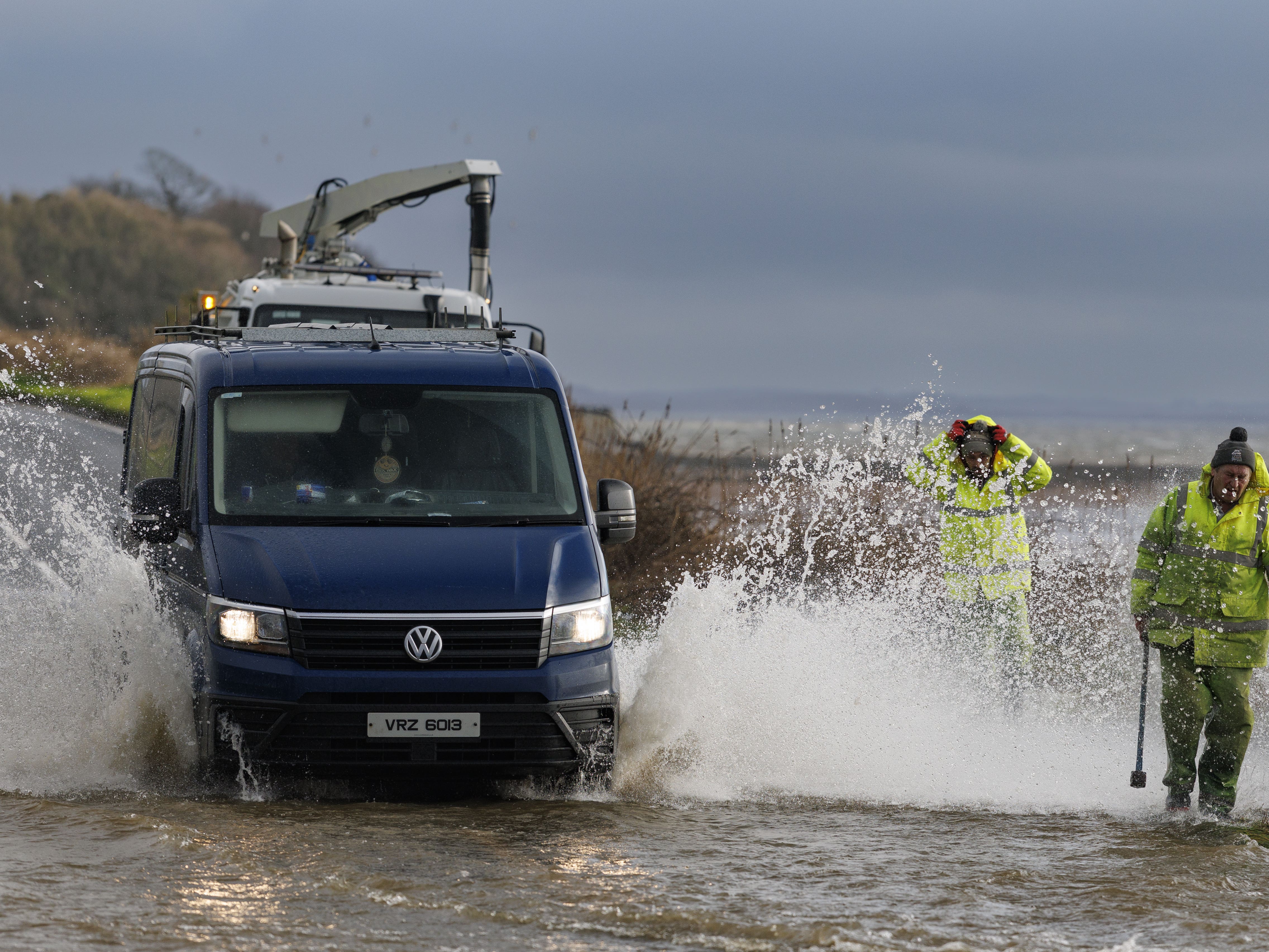 A van is driven through floodwater, splashing two men in hi-vis waterproofs walking nearby
